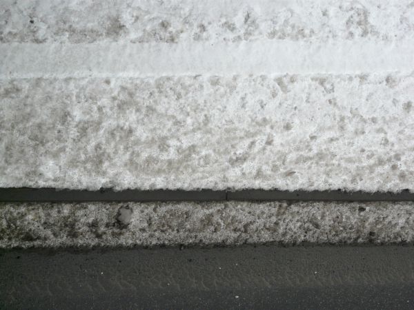 Road edge texture, with grey asphalt embedded with white rocks meeting edges of black-flecked snow, grey sidewalk and a white snow with grey patches.